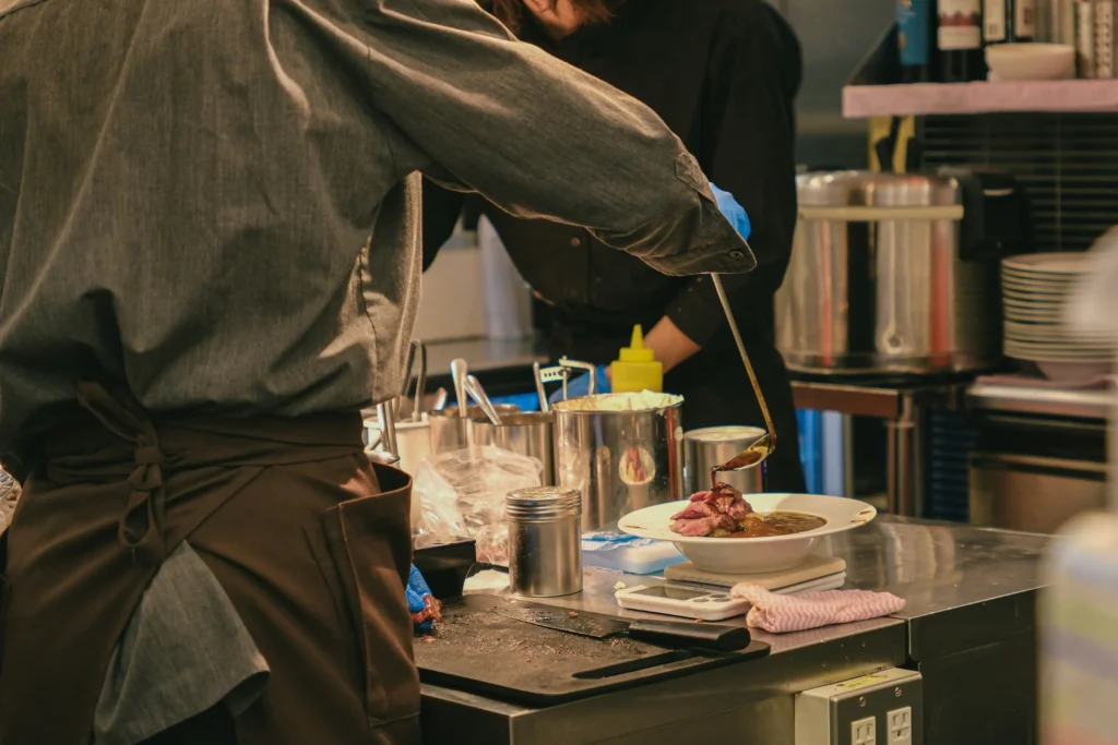 In the image, a chef in a grey shirt and brown apron carefully ladles a dark sauce over a plated dish of meat. The scene captures a busy, warm-toned professional kitchen filled with stainless steel prep stations and various culinary tools.