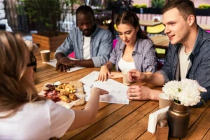 In the image, a group of friends is gathered around a wooden table at an outdoor cafe. They are looking over menus and sharing a platter of finger foods like french fries and fried chicken while deciding what to order.
