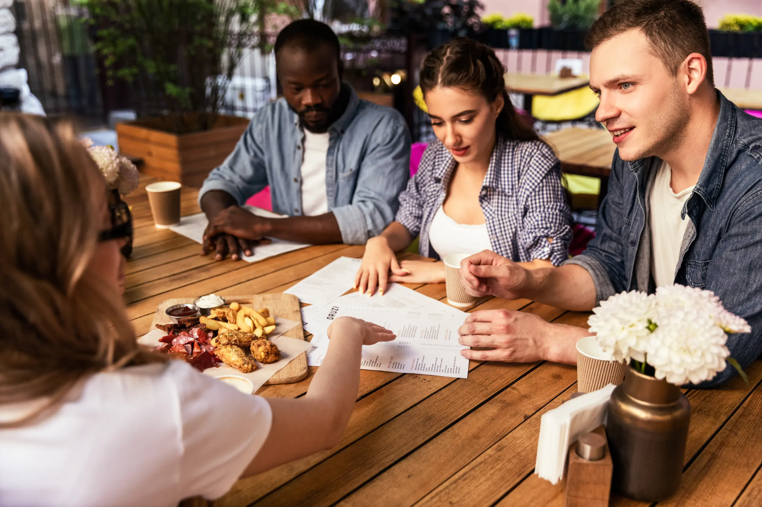 In the image, a group of friends is gathered around a wooden table at an outdoor cafe. They are looking over menus and sharing a platter of finger foods like french fries and fried chicken while deciding what to order.