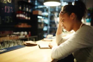 A woman wearing an apron sits at a bar counter with her head resting on her hand, looking down at an open notebook with a stressed or tired expression. The background shows a dimly lit bar or restaurant setting with shelves of bottles and a chalkboard.