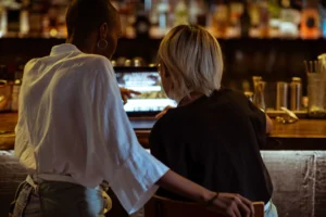 In the image, two people are viewed from behind at a dimly lit bar as they look at an open laptop screen together. One person in a white shirt points at the screen while resting a hand on the back of the other person's chair.