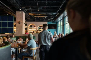 A view from behind a foreground figure looks into a busy, modern restaurant filled with patrons seated at tables. In the center, a waiter stands with his back to the camera, looking toward tables near a large window and a bar with arched entryways in the background.