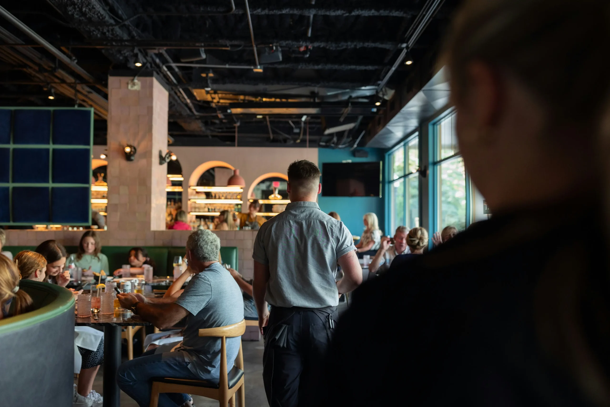 A view from behind a foreground figure looks into a busy, modern restaurant filled with patrons seated at tables. In the center, a waiter stands with his back to the camera, looking toward tables near a large window and a bar with arched entryways in the background.