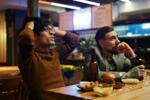 In the image, two men sit at a bar counter with food and drinks, looking intensely toward something out of frame. One man in a brown sweater holds his hands to his head in apparent frustration or disbelief, while the other with tattoos watches intently with a tense expression.