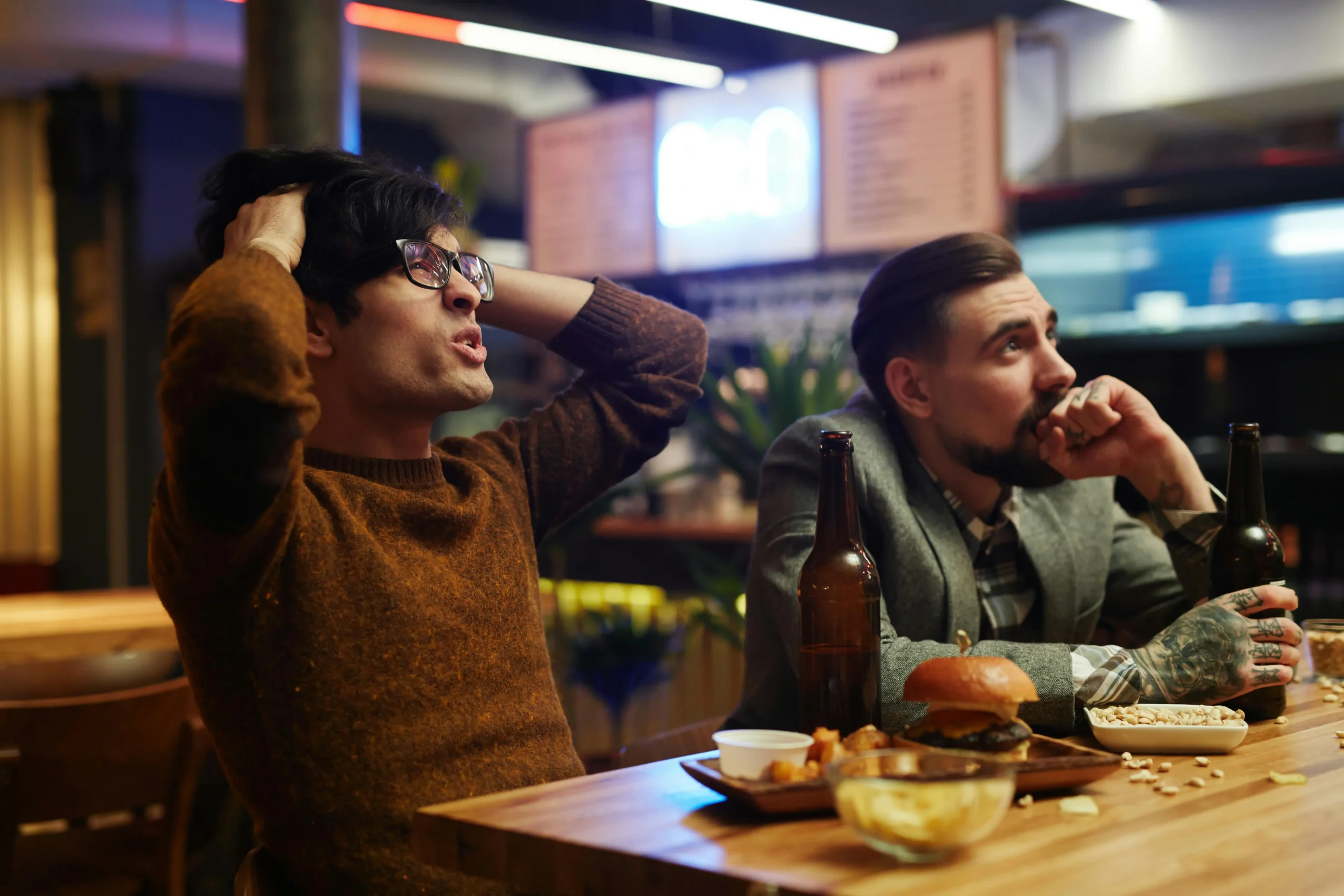 In the image, two men sit at a bar counter with food and drinks, looking intensely toward something out of frame. One man in a brown sweater holds his hands to his head in apparent frustration or disbelief, while the other with tattoos watches intently with a tense expression.
