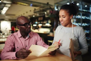 In the image, a man wearing glasses and a pink button-down shirt sits at a restaurant table while reviewing a menu. A smiling waitress wearing an apron stands beside him, holding a notebook and pencil as she prepares to take his order.