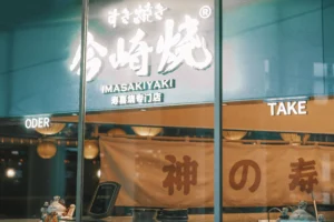 A brightly lit storefront window features a large glowing sign that reads "IMASAKIYAKI" in English, accompanied by Japanese hiragana and Chinese characters. Below the sign, indoor order and takeout windows are visible alongside a large fabric banner and decorative lucky cat figurines.