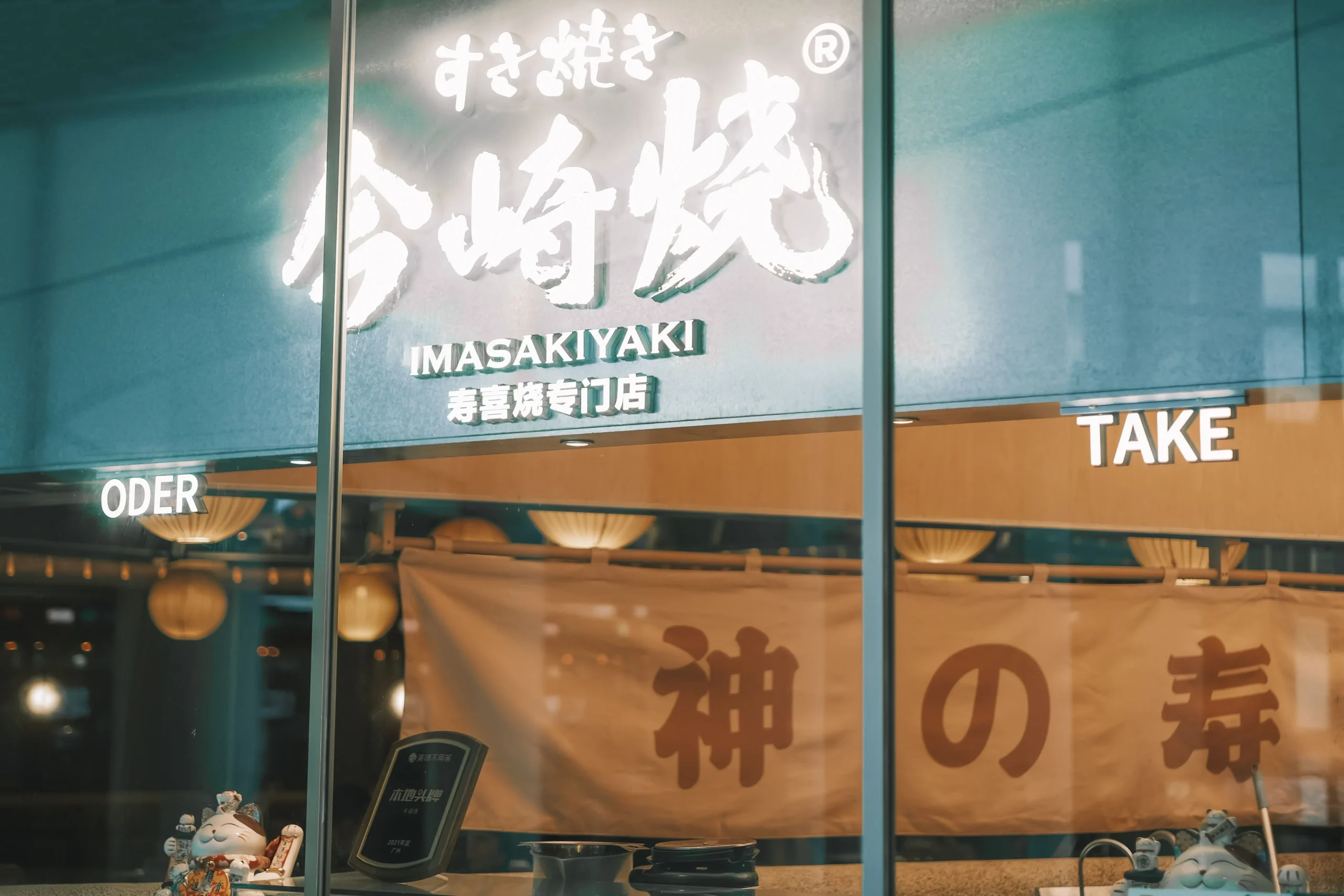 A brightly lit storefront window features a large glowing sign that reads "IMASAKIYAKI" in English, accompanied by Japanese hiragana and Chinese characters. Below the sign, indoor order and takeout windows are visible alongside a large fabric banner and decorative lucky cat figurines.