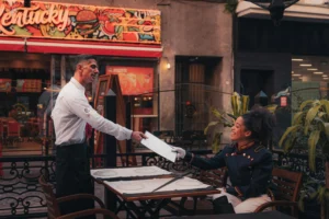In the image, a waiter in a white shirt and black apron hands a menu to a smiling woman seated at an outdoor restaurant table. The woman is dressed in an ornate, dark military-style jacket, and a prop sword rests on the table between them in front of a colorful storefront.