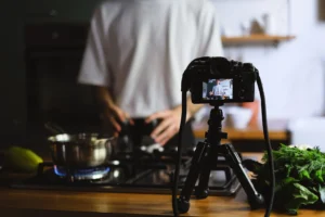 A digital camera mounted on a small tripod records a person in a white t-shirt cooking in a kitchen. In the foreground, a pot heats on a lit gas stove burner next to a bundle of fresh green herbs.