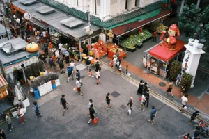 An overhead shot captures a bustling, pedestrian-only street intersection in a vibrant Chinatown district lined with outdoor market stalls selling fruits and merchandise. Numerous people, many wearing face masks, stroll through the paved street while a large, inflatable Caishen (Chinese God of Wealth) figure stands prominently on a red-canopied booth.