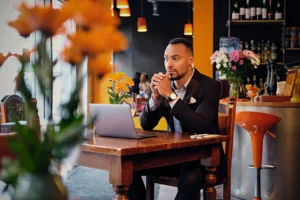 A well-dressed Black man in a dark suit sits at a wooden table in a cafe, looking thoughtful with his hands clasped near his chin while working on a laptop. Orange flowers in the foreground and a bar area in the background create a warm, softly blurred setting around him.
