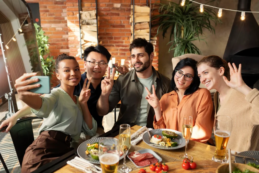 In the image, a diverse group of five friends sits closely together at a wooden dining table filled with food and drinks, smiling warmly for a selfie. A woman on the left holds up a smartphone to capture the group, while several friends flash peace signs and wave toward the camera in a cozy, rustic restaurant setting.