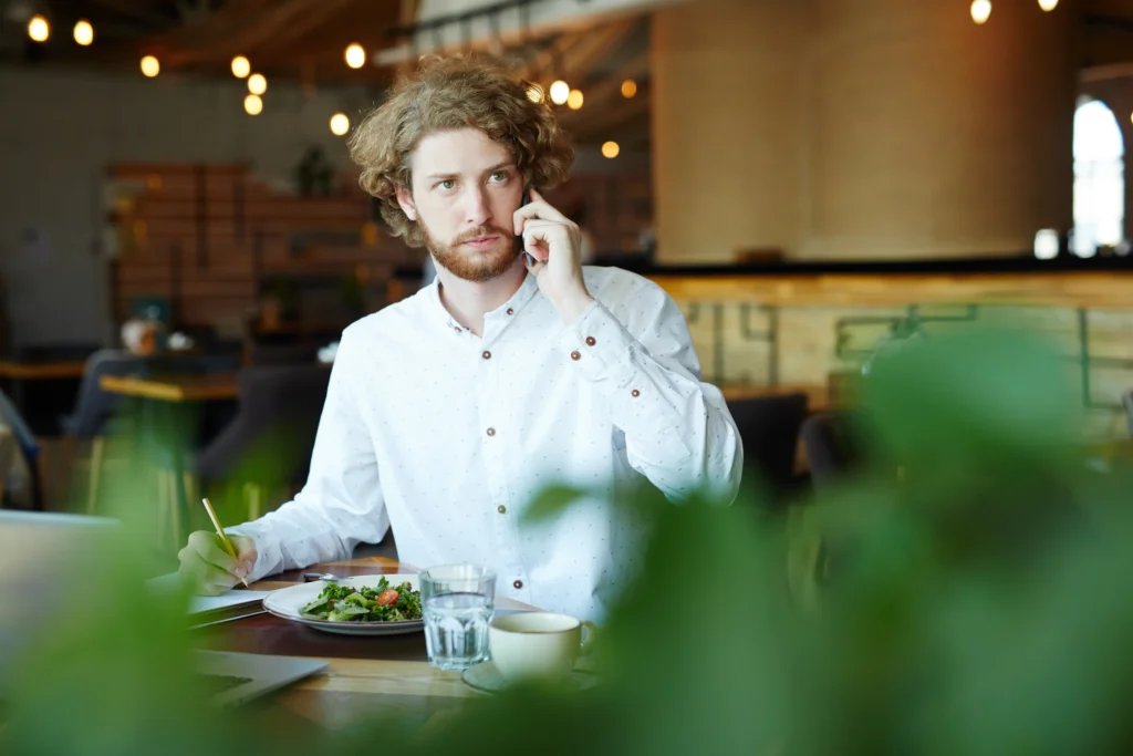A young man with curly hair and a beard talks on his smartphone while sitting at a restaurant table. He holds a pencil ready to take notes next to his meal, laptop, and drinks.