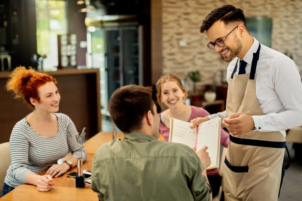A smiling waiter wearing glasses and a beige apron shows a menu to a group of three young adults seated at a wooden restaurant table. The patrons, including a man with his back to the camera and two smiling women, look engaged in the friendly interaction.