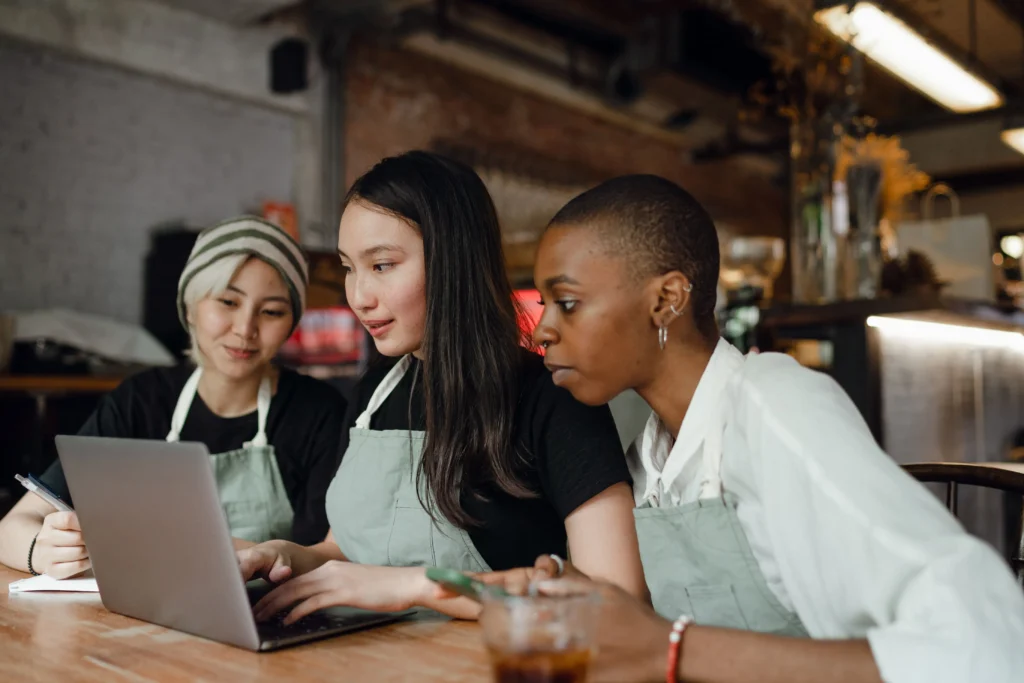 Three diverse cafe workers wearing light green aprons gather closely around a wooden table to look at a laptop screen. The woman in the center types on the keyboard while her colleagues look on attentively, with a warm, rustic coffee shop interior blurred in the background.