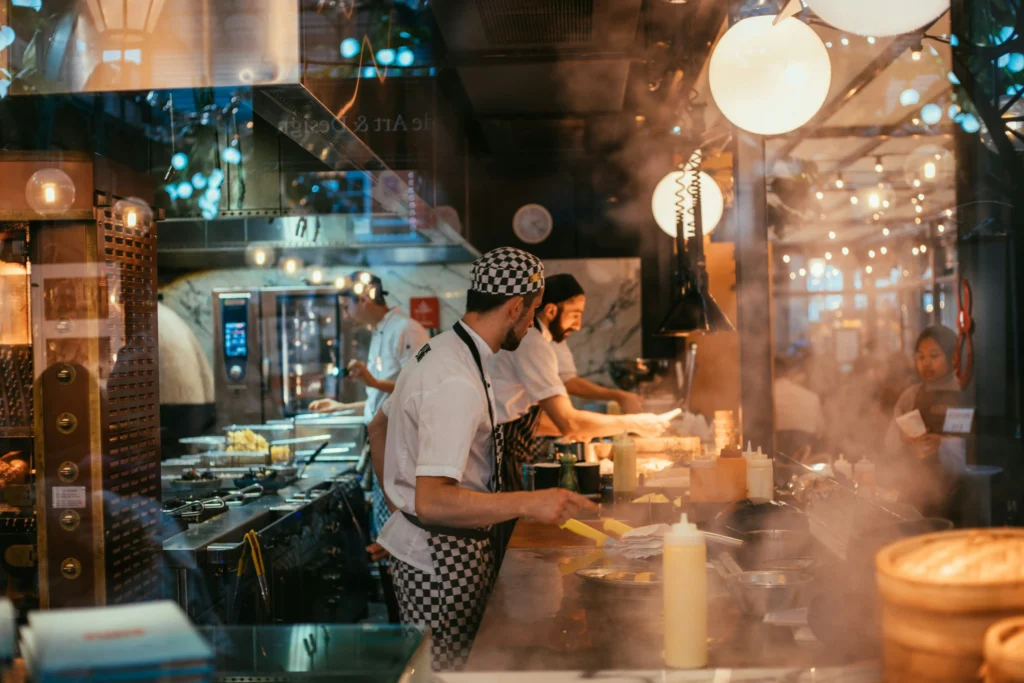 Viewed through a glass window, several chefs wearing white uniforms and patterned aprons work diligently in a busy, warmly lit restaurant kitchen filled with steam. The bustling kitchen line features multiple food preparation stations, stainless steel equipment, and bright, spherical overhead lights.