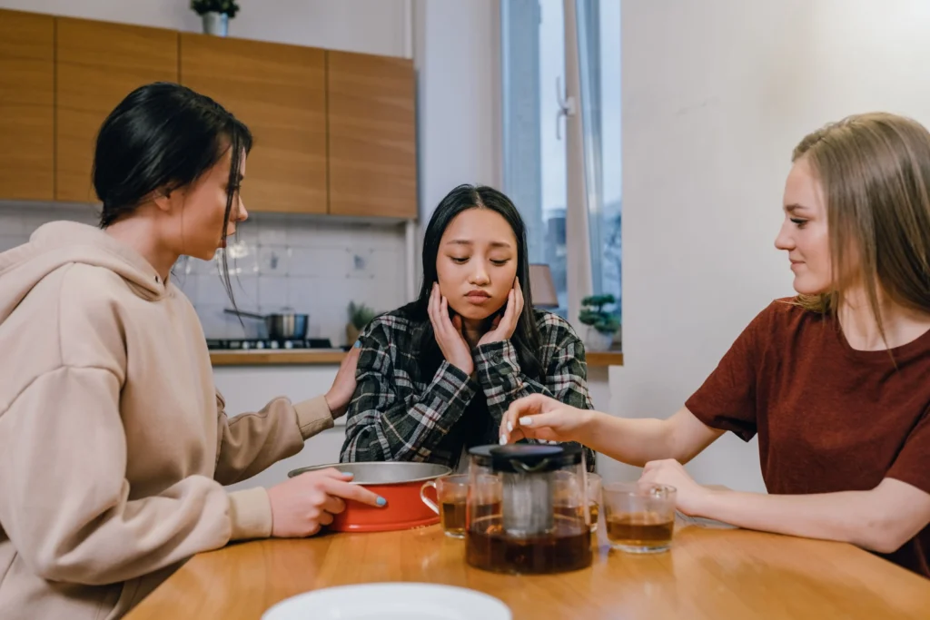 Three young women sit around a wooden kitchen table, where one looks visibly upset while holding her face. Her two friends offer support, with one placing a comforting hand on her shoulder and the other tending to a teapot on the table.