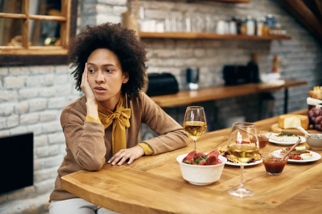 In the image, a woman with an afro hairstyle sits alone at a wooden dining table, leaning her head on her hand with a sad and disappointed expression. The table in front of her is beautifully set with multiple wine glasses, a bowl of strawberries, and various appetizers, suggesting she is waiting for someone who hasn't arrived.