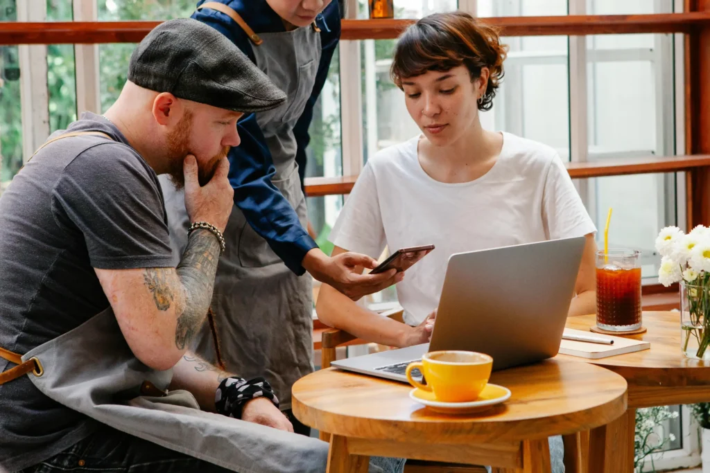 Three people, including a person in an apron holding a smartphone, gather around a laptop at a wooden table in a cafe. A yellow coffee cup sits in the foreground, while a glass of iced coffee and a notebook rest on a nearby table.