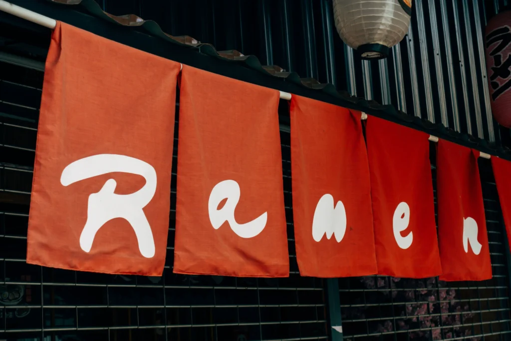 A series of bright red, fabric *noren* curtains hang in a row outside a shop storefront, spellling out the word "Ramen" in stylized white English letters. In the background, a dark ribbed wall and traditional Japanese paper lanterns are visible above a closed security gate.