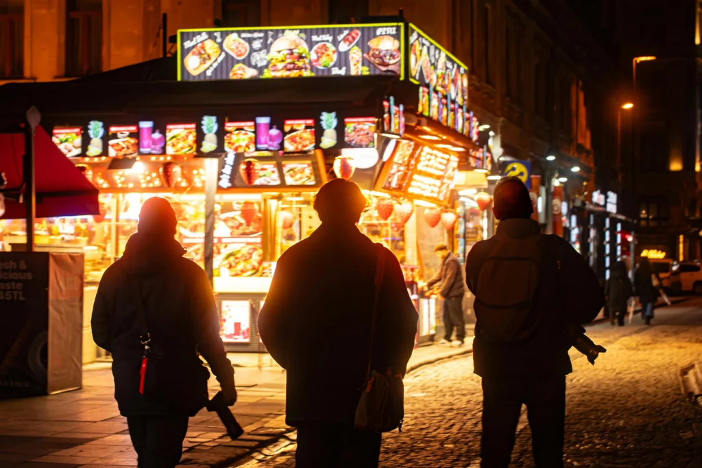 Three people are seen from behind as silhouettes walking down a cobblestone street at night toward a brightly lit, glowing fast-food stand. The vibrant food stall features colorful illuminated menus displaying burgers, wraps, and drinks, casting a warm orange glow over the street.
