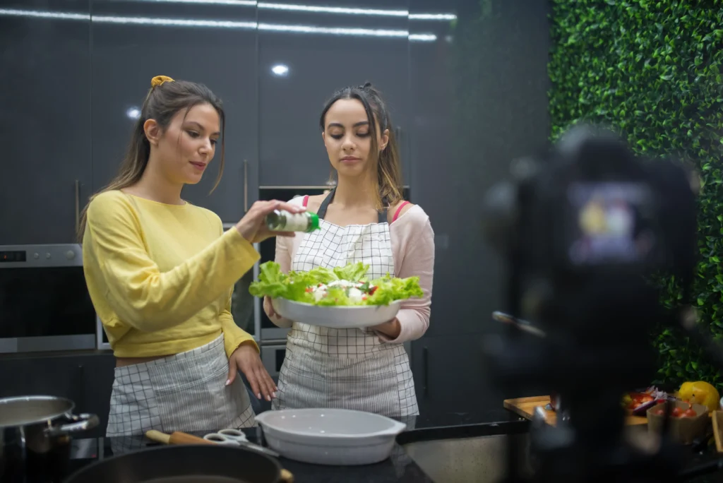 In the image, two young women are preparing a fresh green salad together in a modern dark-gray kitchen. One woman in a pink top and grid-patterned apron holds a large bowl of salad while her companion in a yellow sweater seasons it, all being recorded by a camera blurred in the foreground.