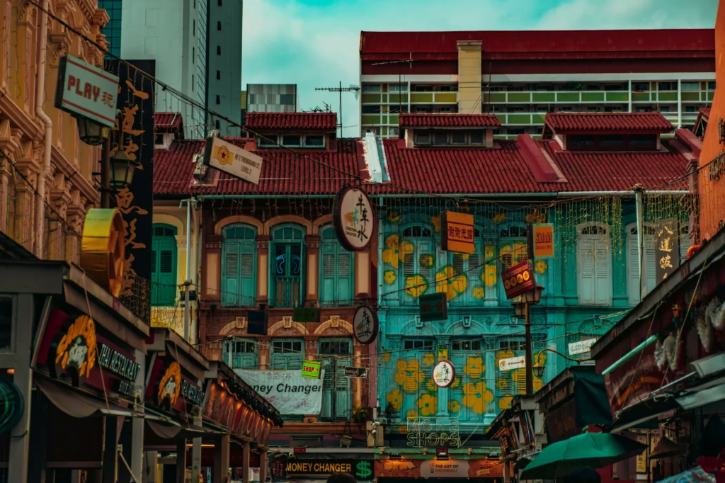 A vibrant street scene in Singapore's Chinatown features historic, multi-story shophouses decorated with intricate facades, colorful shutters, and hanging signs. The foreground is bustling with storefronts, including a money changer and a restaurant, set against a backdrop of modern high-rise buildings.
