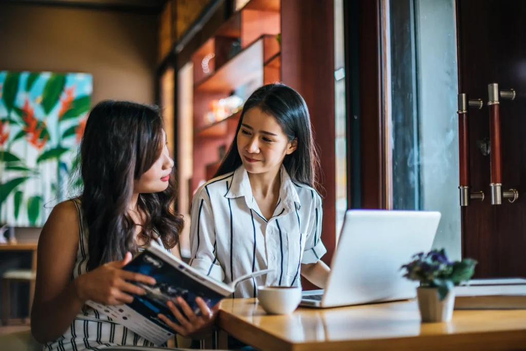 Two women sit at a wooden table inside a cafe, engaged in a conversation with a laptop and a coffee cup in front of them. One woman holds open a magazine while the other looks at her responsively against a backdrop of warm lighting and shelves.