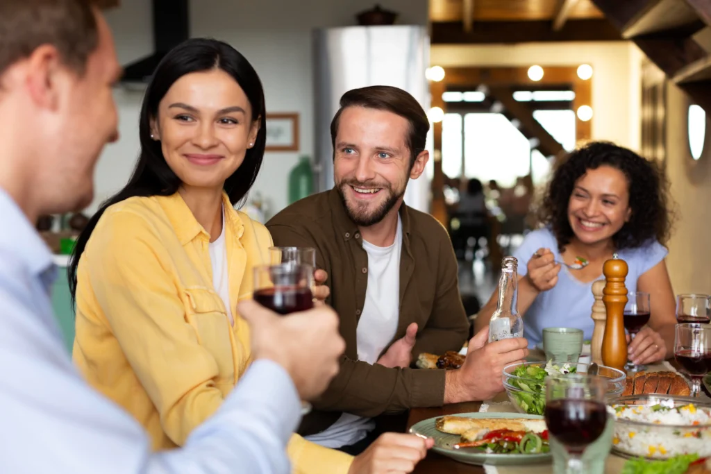 A group of friends are smiling and laughing together while sharing a meal around a dining table. They are holding up drinks, chatting, and enjoying a variety of food including salads, rice, and bread in a warmly lit home setting.