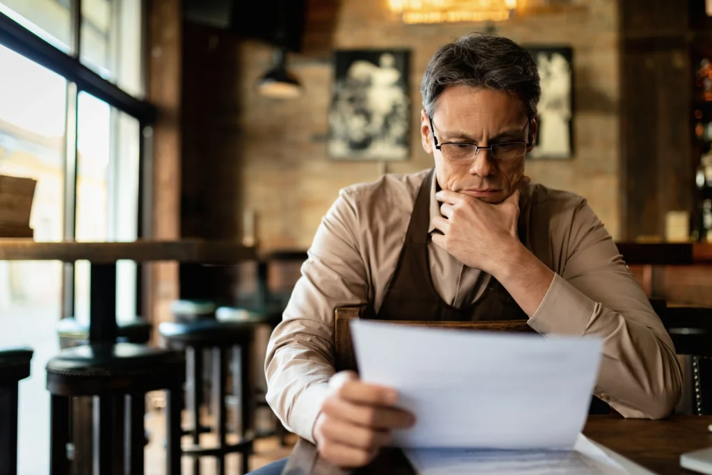 A pensive man wearing glasses and a barista apron sits in a dimly lit café, reviewing a document. He holds the paper in one hand while resting his chin on the other with a concentrated expression.