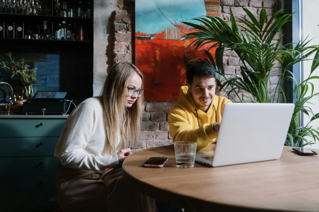 In the image, a man in a yellow hoodie and a woman wearing glasses sit together at a wooden table, focused on an open laptop screen. They are in a rustic cafe or office setting featuring an exposed brick wall, a vibrant abstract painting, and a large potted plant.