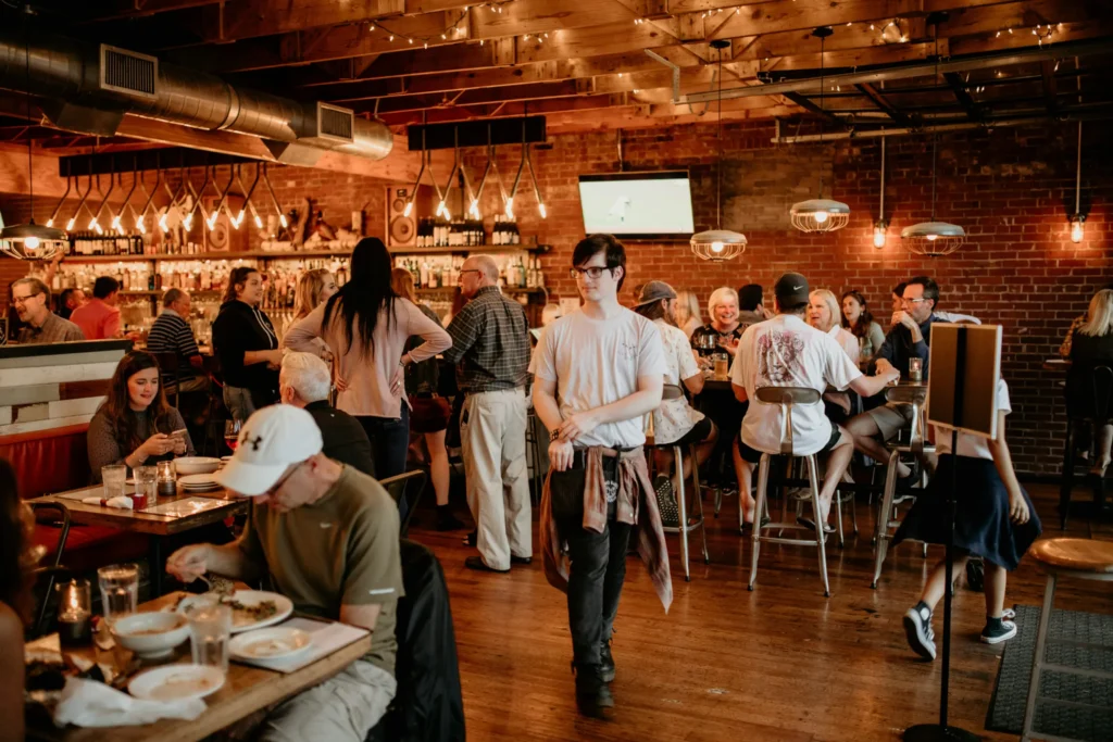 A bustling restaurant interior features a waiter walking through the center of a crowded dining room with exposed brick walls and warm, hanging lights. Numerous patrons are seated at tables and bars, talking and eating in a lively, rustic atmosphere.