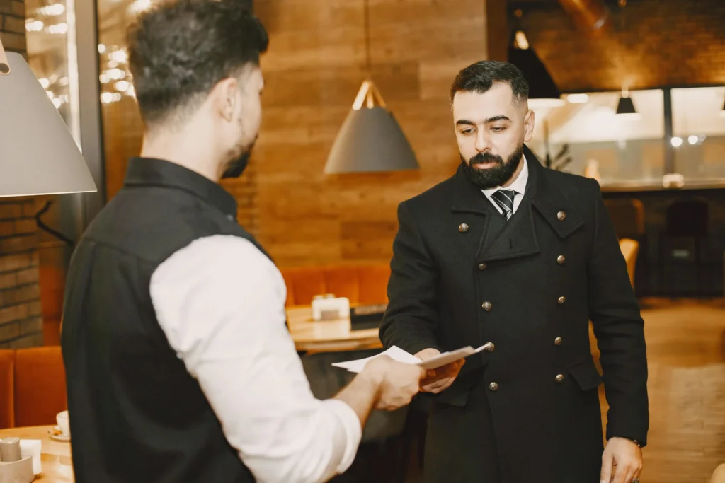 In the image, a man with a beard wearing a dark, double-breasted coat and tie is handing papers to a restaurant server or host. The background features a warmly lit, modern restaurant interior with wooden walls and hanging lamps.