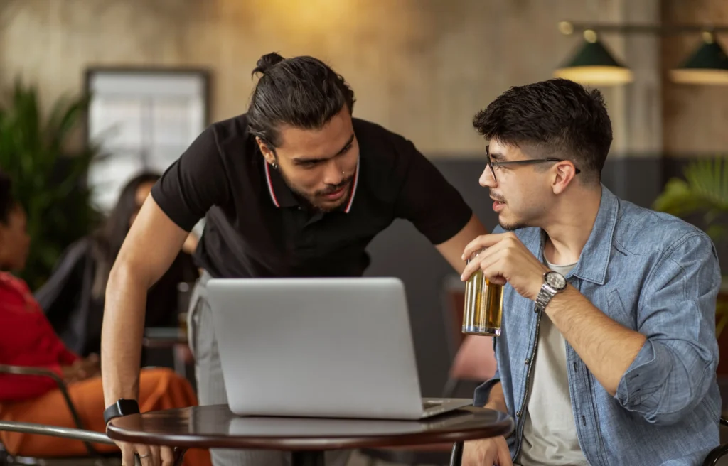 Two men are conversing over a laptop at a small table in a dimly lit cafe or workspace. One man leans in from the left wearing a black polo shirt, while the other sits on the right in a denim shirt, holding a glass of a beverage.