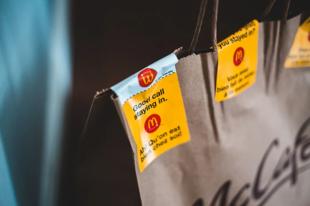 A close-up shot shows a brown McCafé paper delivery bag sealed with a bright yellow McDonald's sticker. The sticker feature the brand's logo alongside the bilingual text, "Good call staying in" and "Ah! Qu'on est bien chez soi!"