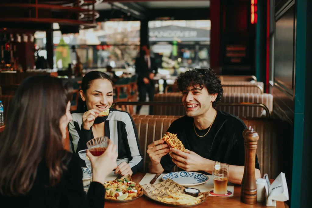 Three friends laugh and chat while enjoying a meal together at a restaurant booth. The table is filled with food, including nachos, French fries, and a hot dog, alongside refreshing drinks.