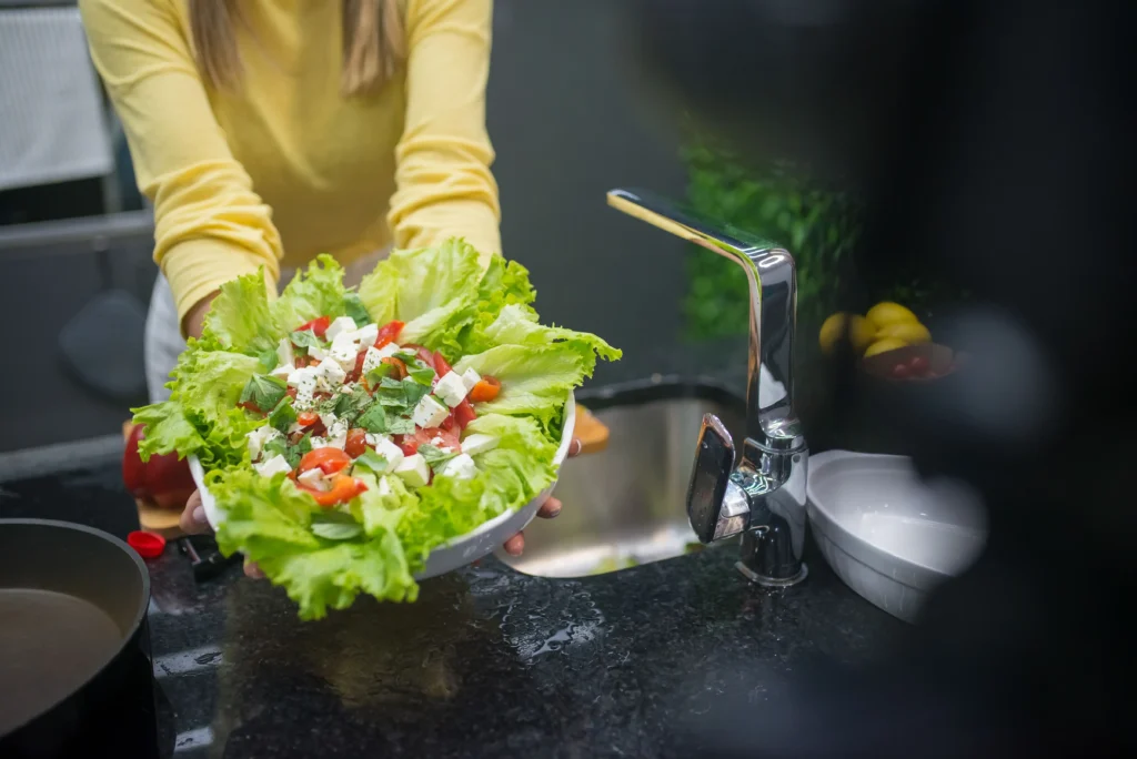 A person in a yellow long-sleeved shirt holds out a large white bowl filled with a vibrant green salad topped with cubed feta cheese and tomatoes. The scene is set in a kitchen, with a modern chrome faucet and a kitchen sink visible in the background.