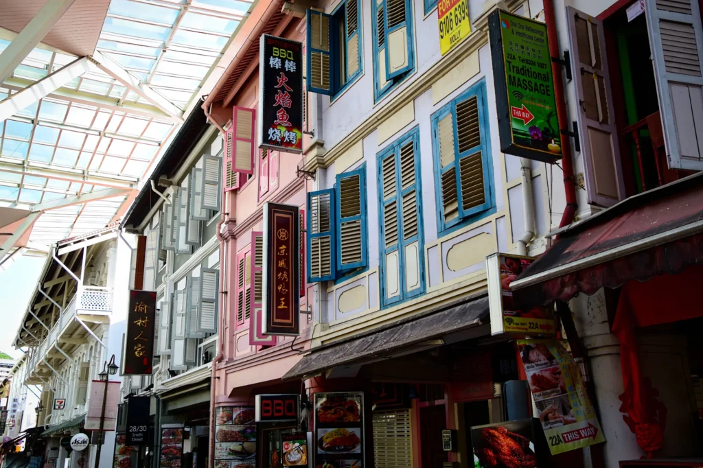 A low-angle shot showcases a row of vibrant, multi-story heritage shophouses featuring colorful wooden shutters and numerous prominent business signs in English and Chinese. The streetscape is sheltered by a modern, overhead glass and metal canopy that allows natural light to filter down.