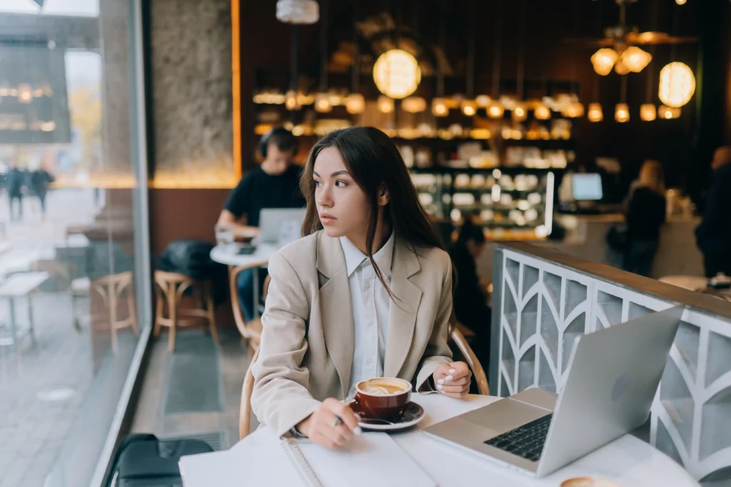 A young woman in a beige blazer sits at a cafe table with a laptop, a notebook, and a cup of coffee while looking out the window. In the blurred background, other patrons work and warm decorative lights illuminate the cozy interior.