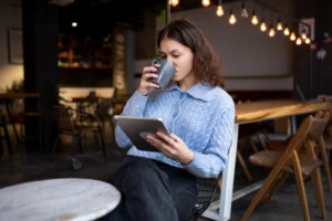 A young woman sitting in a cozy, warm-lit cafe sips a drink from a grey mug while holding a tablet. She wears a light blue knit cardigan and appears focused on reading or working on her device.