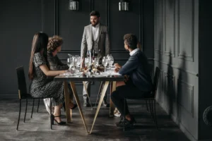 In the image, a well-dressed man in a grey suit stands at the head of a formal dinner table, smiling as he addresses a group of seated guests. The dinner party takes place in an elegant room with dark paneled walls, featuring a beautifully set table filled with wine glasses and food.