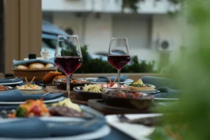 Two wine glasses filled with red wine stand on an outdoor restaurant table laden with various small plates, appetizers, and main dishes. The background shows a soft-focus street view, creating an inviting and warm dining atmosphere.