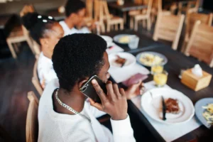 A young man wearing a white shirt and silver chain talks on his smartphone while sitting at a restaurant dining table. He is surrounded by friends, plates of food, and drinks during a group meal.