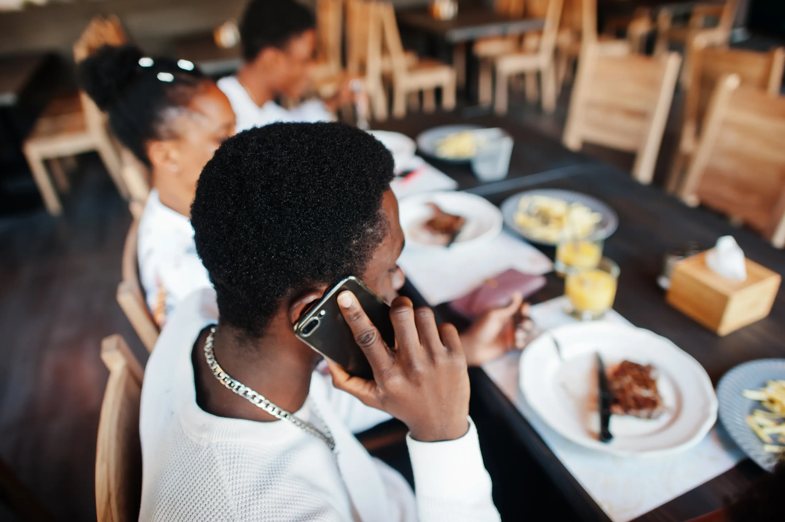 A young man wearing a white shirt and silver chain talks on his smartphone while sitting at a restaurant dining table. He is surrounded by friends, plates of food, and drinks during a group meal.