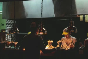 Two kitchen staff members work behind a restaurant counter illuminated by warm hanging heat lamps. Paper cones of french fries sit ready under the light as the worker in the background prepares orders in a cap and apron.