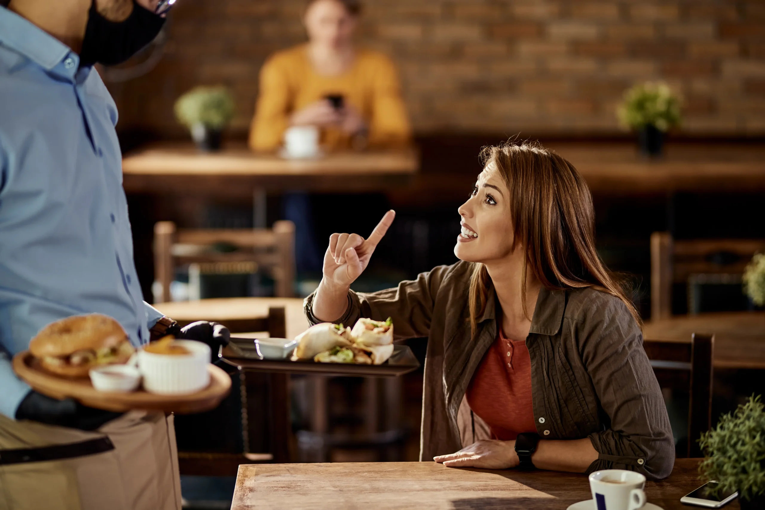 In the image, a young woman sits at a restaurant table and raises her index finger while speaking to a waiter who is holding a tray of wraps and a burger. The waiter wears a face mask and black gloves, while another customer sits blurred in the background of the warm, brick-walled pub.