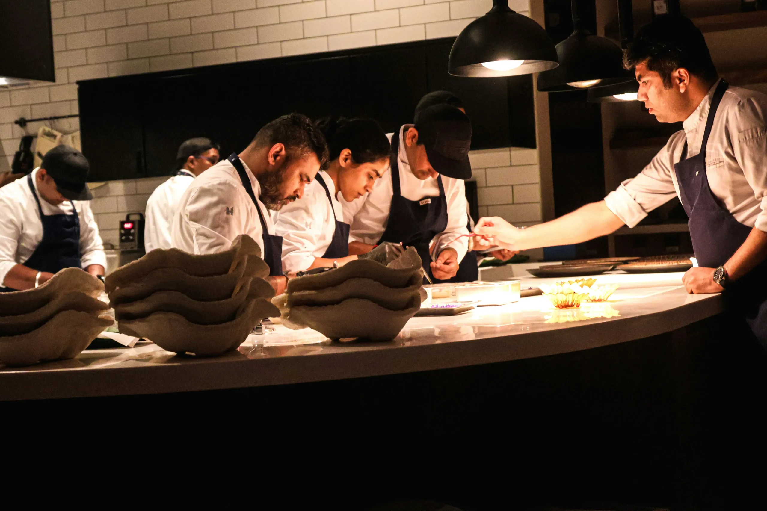 A team of professional chefs in white coats and dark aprons works carefully along a curved kitchen counter under warm, focused overhead lighting. They are intently focused on plating dishes, with stacked artisan ceramic bowls in the foreground and dark cabinetry in the background.