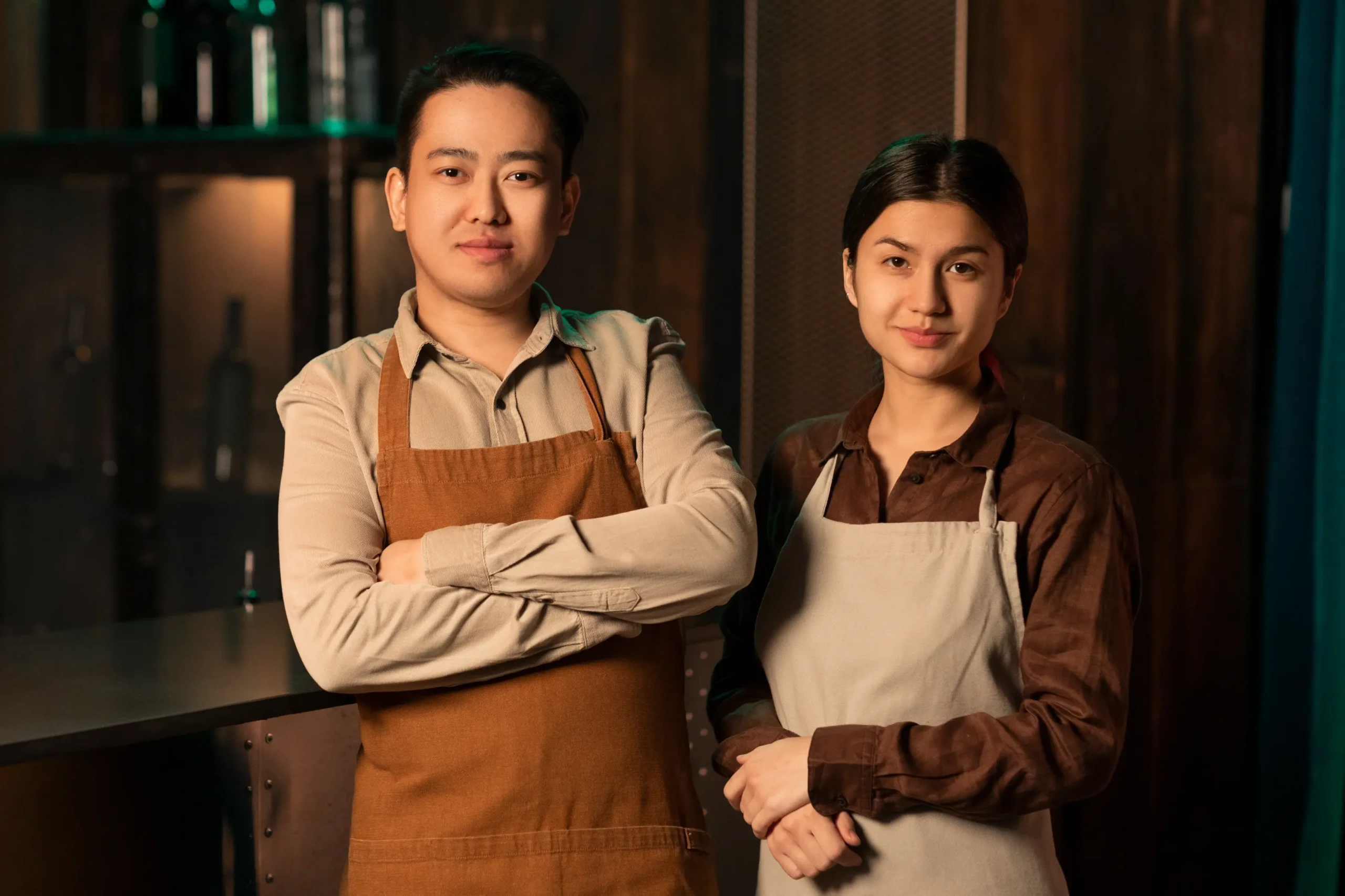 A man with crossed arms and a woman stand side-by-side behind a bar counter, both wearing aprons and looking toward the camera. The background features a dimly lit, rustic bar setting with wooden paneling and shelves.