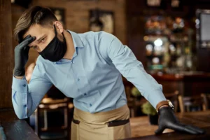 A waiter wearing a black face mask and protective black gloves rests his head on his hand while leaning against a counter, looking visibly stressed and exhausted. He is dressed in a light blue dress shirt and a tan apron inside a dimly lit restaurant or pub setting.